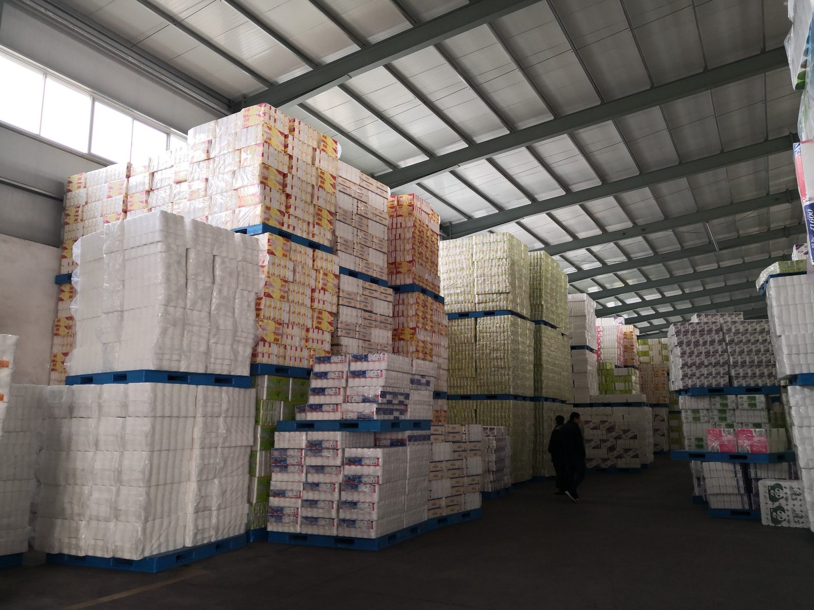 Virgin pulp tissue cartons racked on pallets inside a distribution warehouse, the stacking height and floor clearance that decide how fast stored rolls age